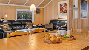 A cozy living room with a wooden ceiling, black leather sofas, a wooden dining table with a basket of bread and fruit, and a framed abstract painting on the wall.