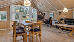 A spacious loft with a high ceiling, wooden floors, a dining area with a table and chairs, a living room with a sofa and a TV, and large windows offering a view of the outdoors.