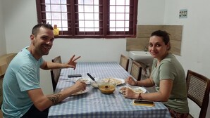 Une table à manger recouverte d’une nappe à carreaux, deux personnes assises, et une fenêtre avec des barreaux.