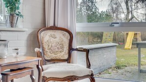 Een kamer met een groot raam, een houten stoel met een bloemendessin, een kleine tafel en een bloempot.