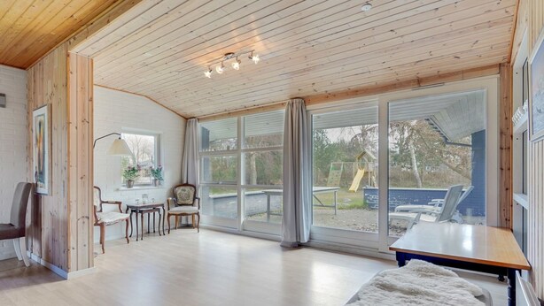 Een ruime kamer met houten vloeren, een groot raam met uitzicht op een speeltuin, een tafel en een pluchen poef.
