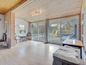Een ruime kamer met houten vloeren, een groot raam met uitzicht op een speeltuin, een tafel en een pluchen poef.