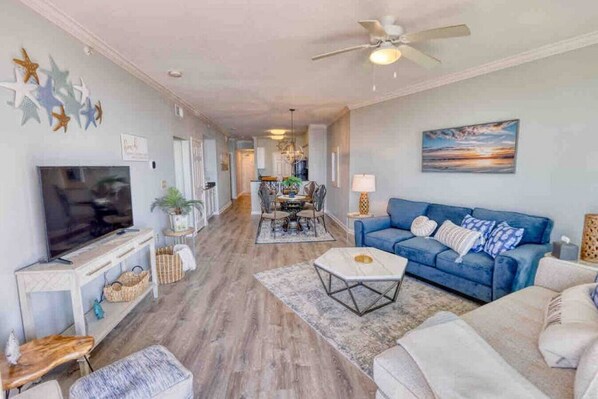 A spacious living room with a television, blue sofa, and a dining area in the background. - Full Ocean View - Gulfport Getaway, 2 Bed/2 Bath (Gulfport)