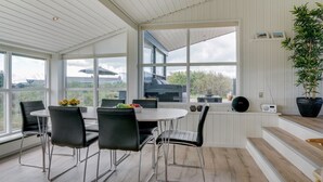 A modern dining area with a large table and chairs, large windows offering views of the outdoors, and a staircase leading to an upper level.