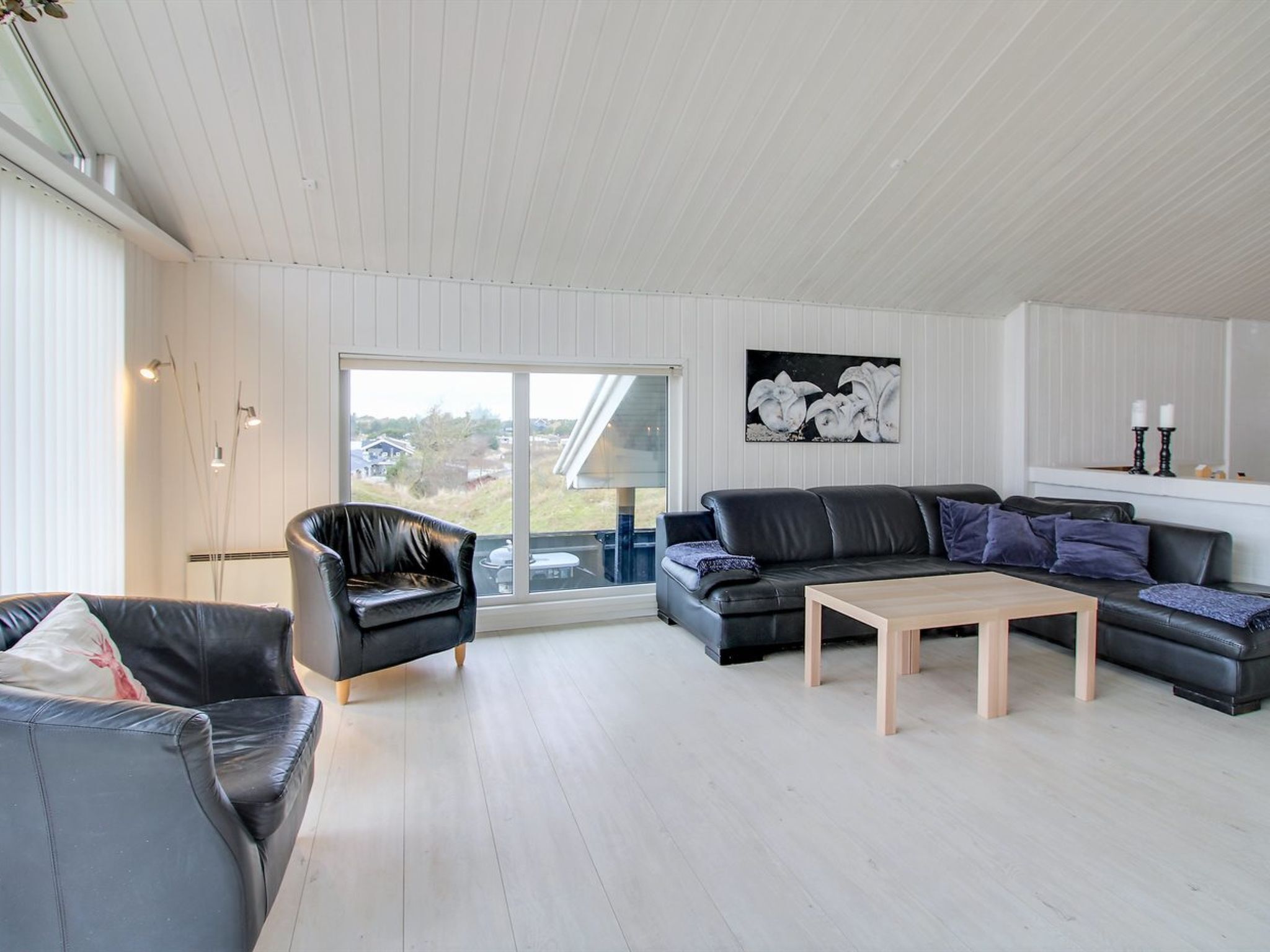 Een moderne woonkamer met een zwarte leren bank, een houten salontafel en een zwarte fauteuil. Een groot raam biedt uitzicht op het landschap.