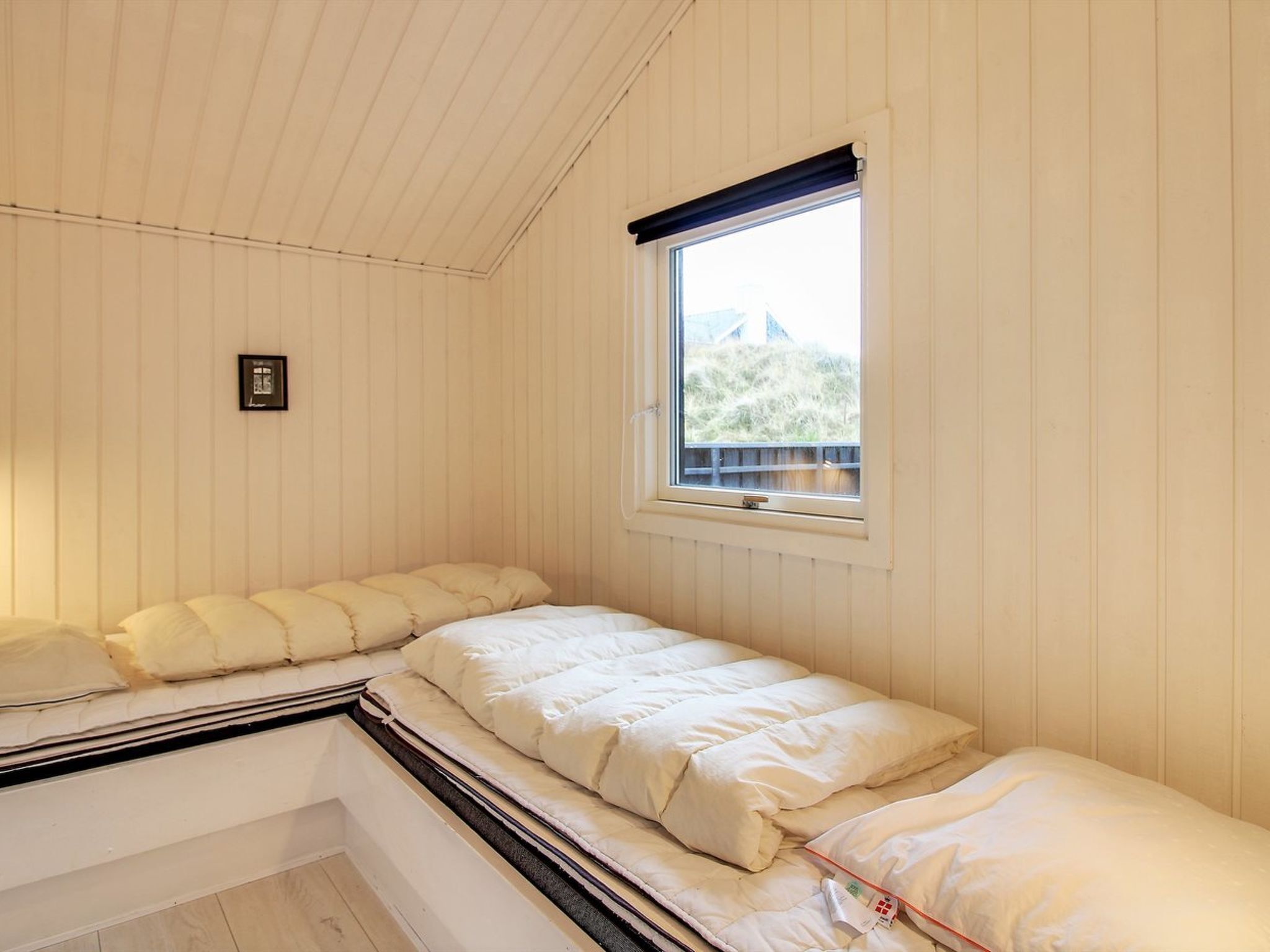 Pequeña habitación con cama individual, ventana, paredes de madera y una cama sencilla con ropa blanca.