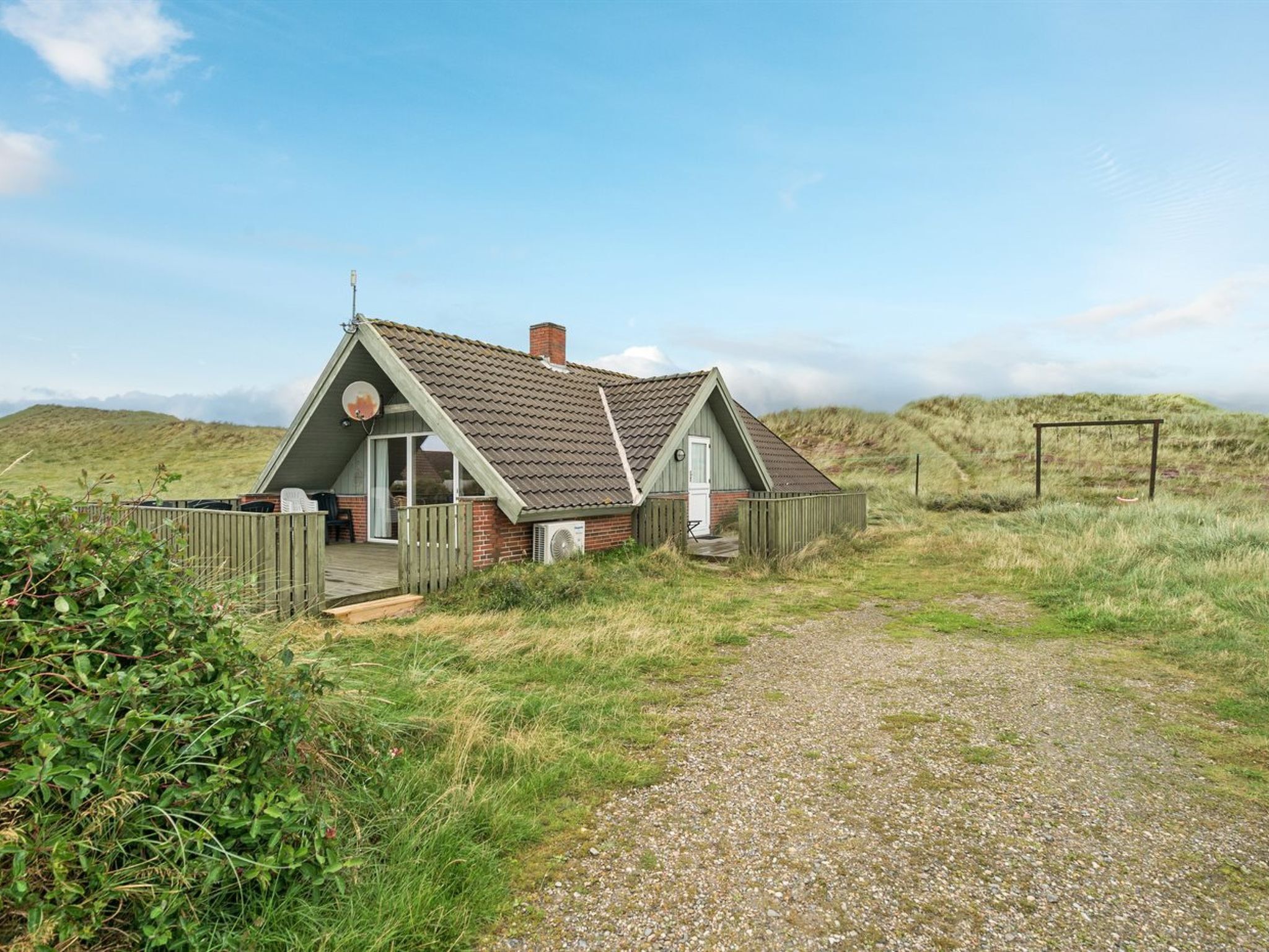 A small, single-story house with a sloped roof, a wooden deck, and a gravel path leading to it.