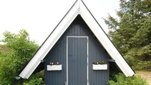 Un petit abri de jardin de couleur sombre, doté d’un toit triangulaire et d’une porte. Deux jardinières blanches sont fixées aux murs extérieurs de l’abri.