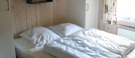 A single bed with white bedding in a room with wooden panel walls, a small shelf, and a window with curtains.