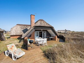 A thatched-roof house with a wooden deck, a barbecue, and a seating area.