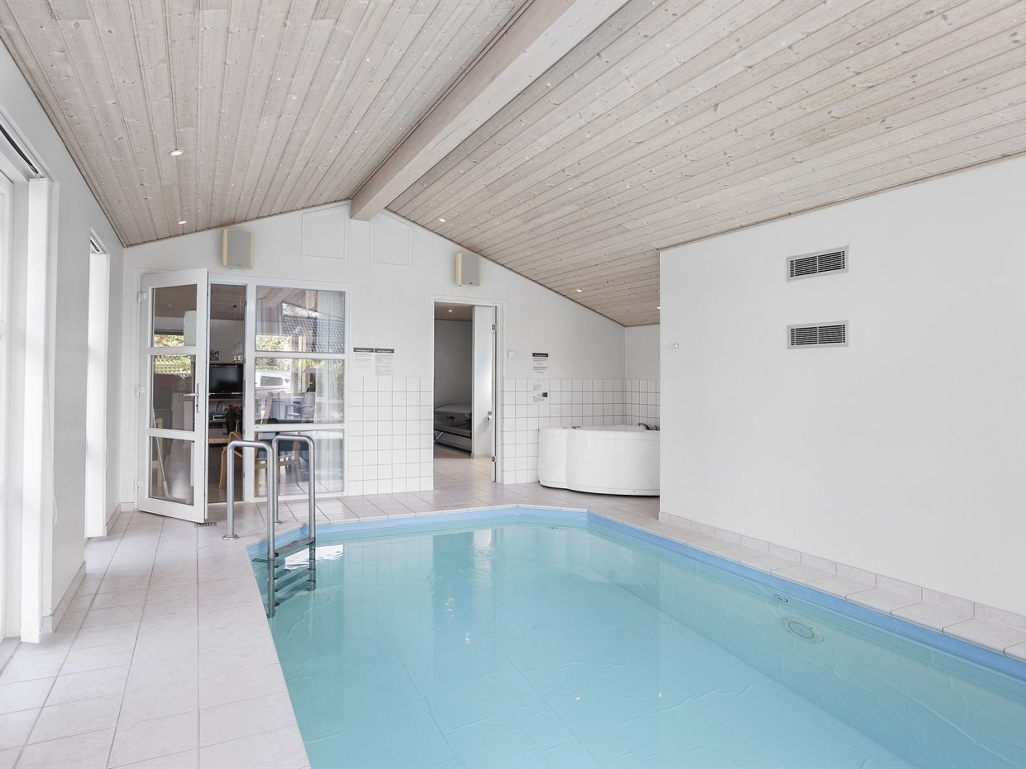 A modern indoor swimming pool with a wooden ceiling, a glass door entrance, and a visible hot tub area.