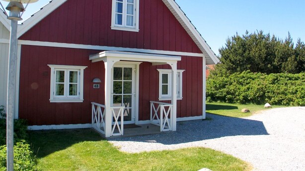 Une maison en bois rouge avec un porche, des boiseries blanches et une plaque portant le numéro « 65 ».