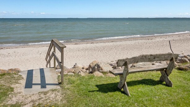 Ein Strand mit einer Holzbank und einer hölzernen Struktur, möglicherweise einem Rettungsschwimmturm, nahe am Wasser.