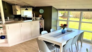 Eine moderne Küche mit weißen Schränken, ein Essbereich mit einem weißen Tisch und grauen Stühlen sowie ein großes Fenster mit Blick ins Freie.