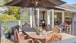 Eine Holzterrasse mit Tisch und Stühlen, einem großen Sonnenschirm und einem gläsernen Anbau mit Kaminofen.