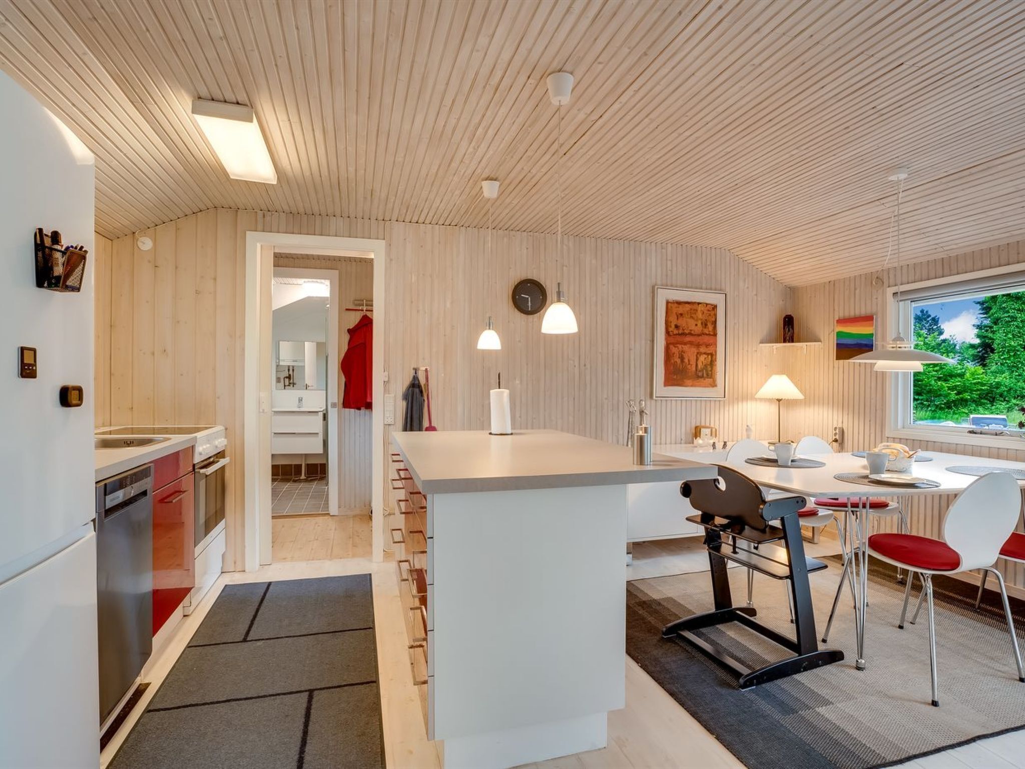 A modern kitchen with a central island, a dining area with a table and chairs, and a view of greenery outside.