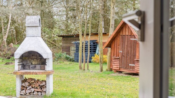 Un four extérieur en pierre, avec un foyer, un abri en bois et une pile de bois de chauffage, le tout situé dans un espace herbeux, avec des arbres en arrière-plan.