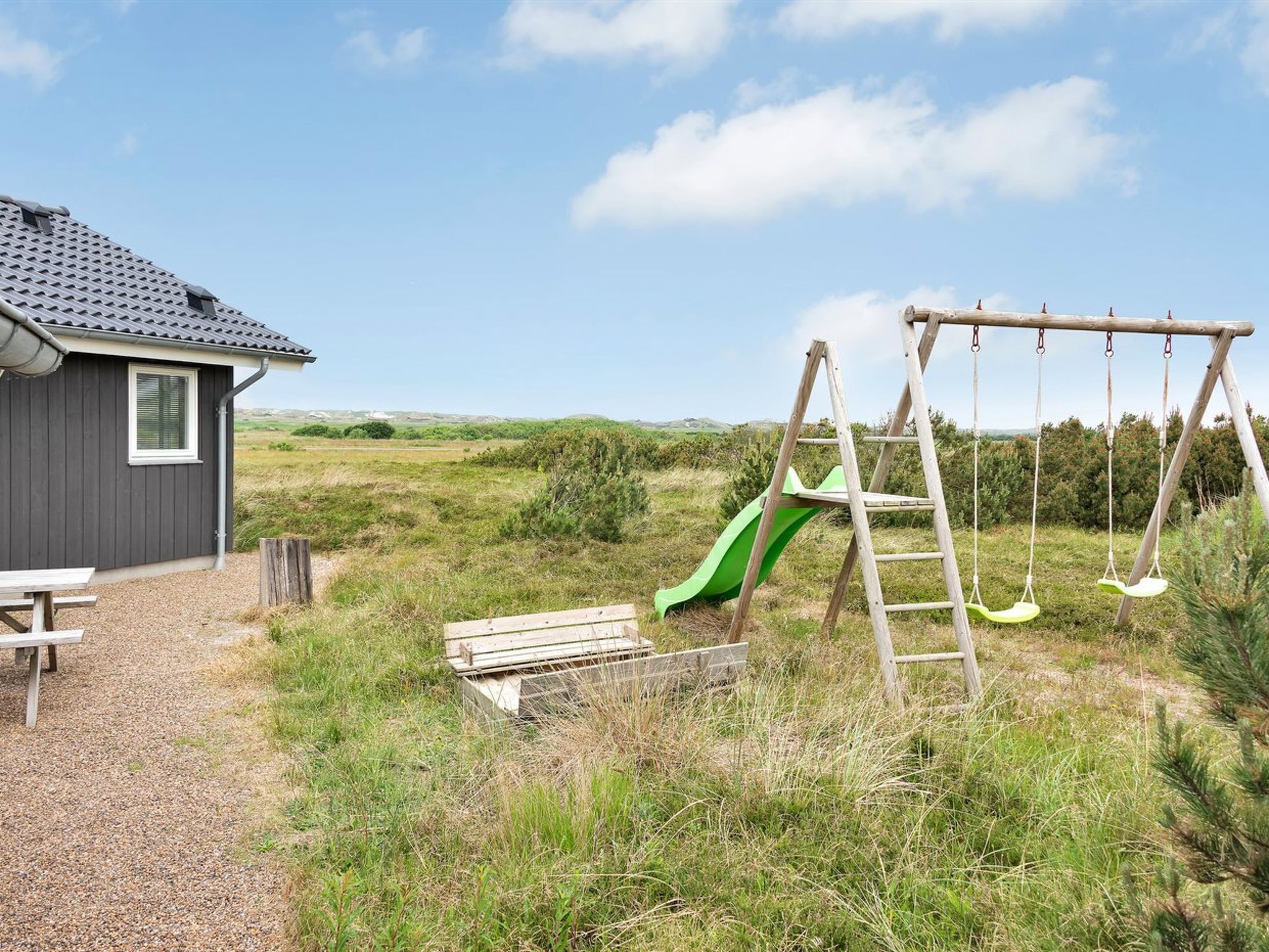 A rustic cabin with a picnic table, a wooden swing set, and a green slide in a grassy field.