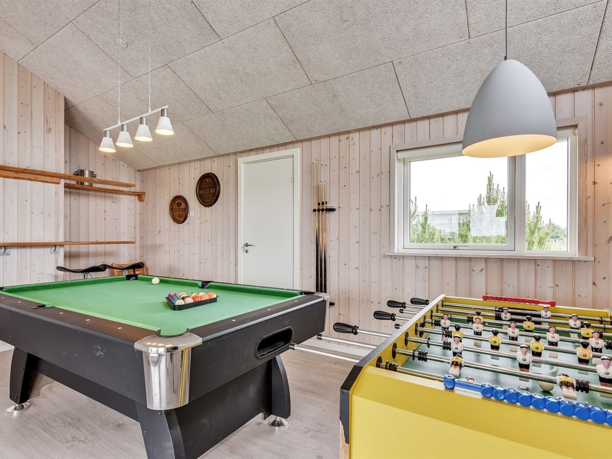 Une pièce équipée d’une table de billard, d’un baby-foot et d’une table de jeu de flipper. Les murs sont en bois et il y a une fenêtre avec vue.