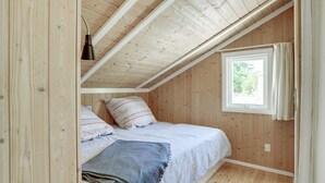 Ein kleines, gemütliches Schlafzimmer im Dachgeschoss mit Holzwänden und schräger Decke. Es gibt ein Einzelbett mit gestreiftem Kopfkissen und einer Decke, ein kleines Fenster mit Vorhängen sowie eine Pendelleuchte.