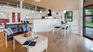 A spacious living area with a wooden ceiling, a dining table with chairs, a sofa with cushions, and a kitchen in the background.