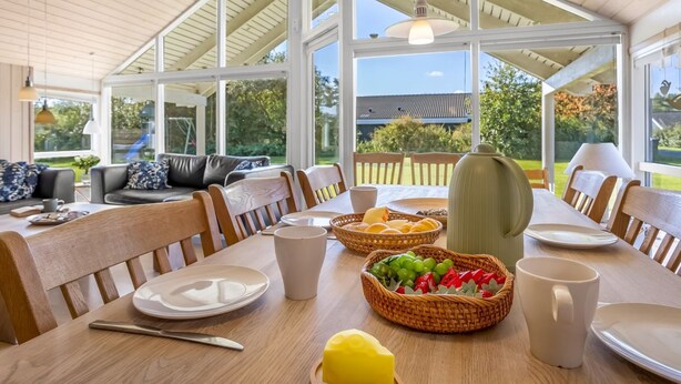 A dining area with a wooden table set for breakfast, surrounded by large windows offering a view of the outdoors. - "Enid" - 700m from the sea by Interhome (Væggerløse)