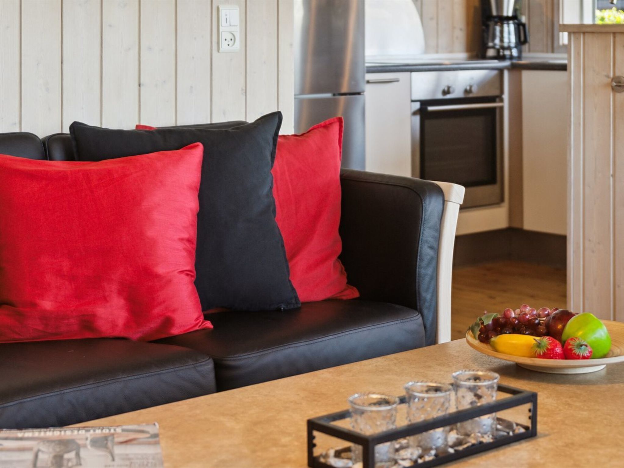 A modern living room with a black leather sofa, red and black cushions, and a wooden dining table with a fruit bowl and glasses.