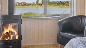 Une pièce chaleureuse avec un poêle à bois, un fauteuil en cuir et une grande fenêtre donnant sur un lac, avec des maisons visibles au loin.