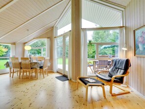 Een ruime kamer met houten vloeren, een eethoek met een tafel en stoelen, een zithoek met een stoel en een bank, en grote ramen met uitzicht op de omgeving.