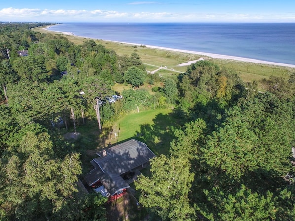 An aerial view of a coastal property with a beach, dense forest, and a house with a red roof.