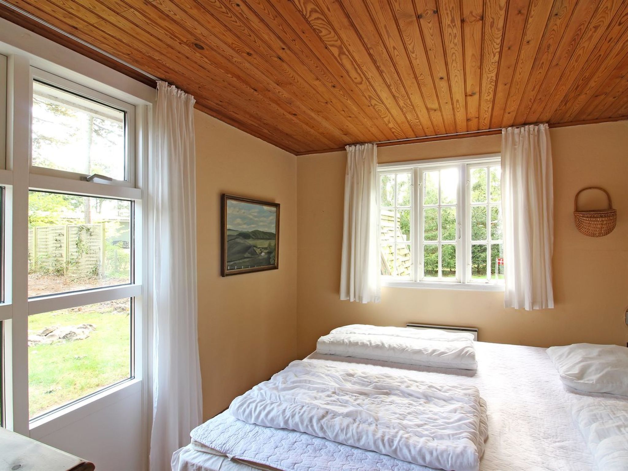 A bedroom with two beds, large windows with white curtains, a wooden ceiling, and a painting on the wall.
