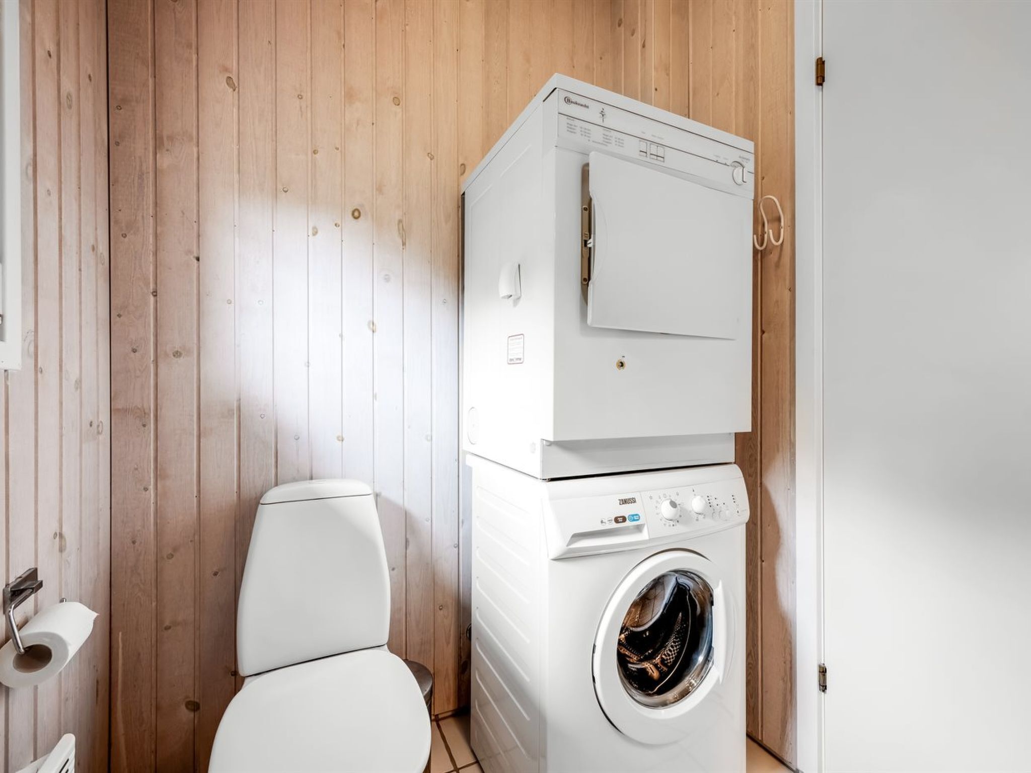 A compact, modern bathroom with wooden walls, a white toilet, and a stacked washer and dryer unit.