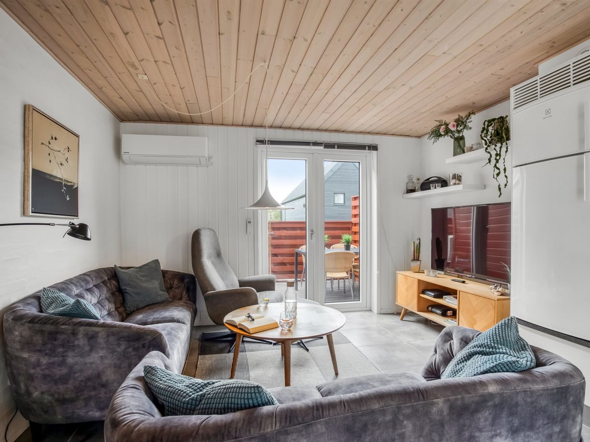 Een moderne woonkamer met een houten plafond, grijze banken, een ronde salontafel en een televisie op een houten kast.