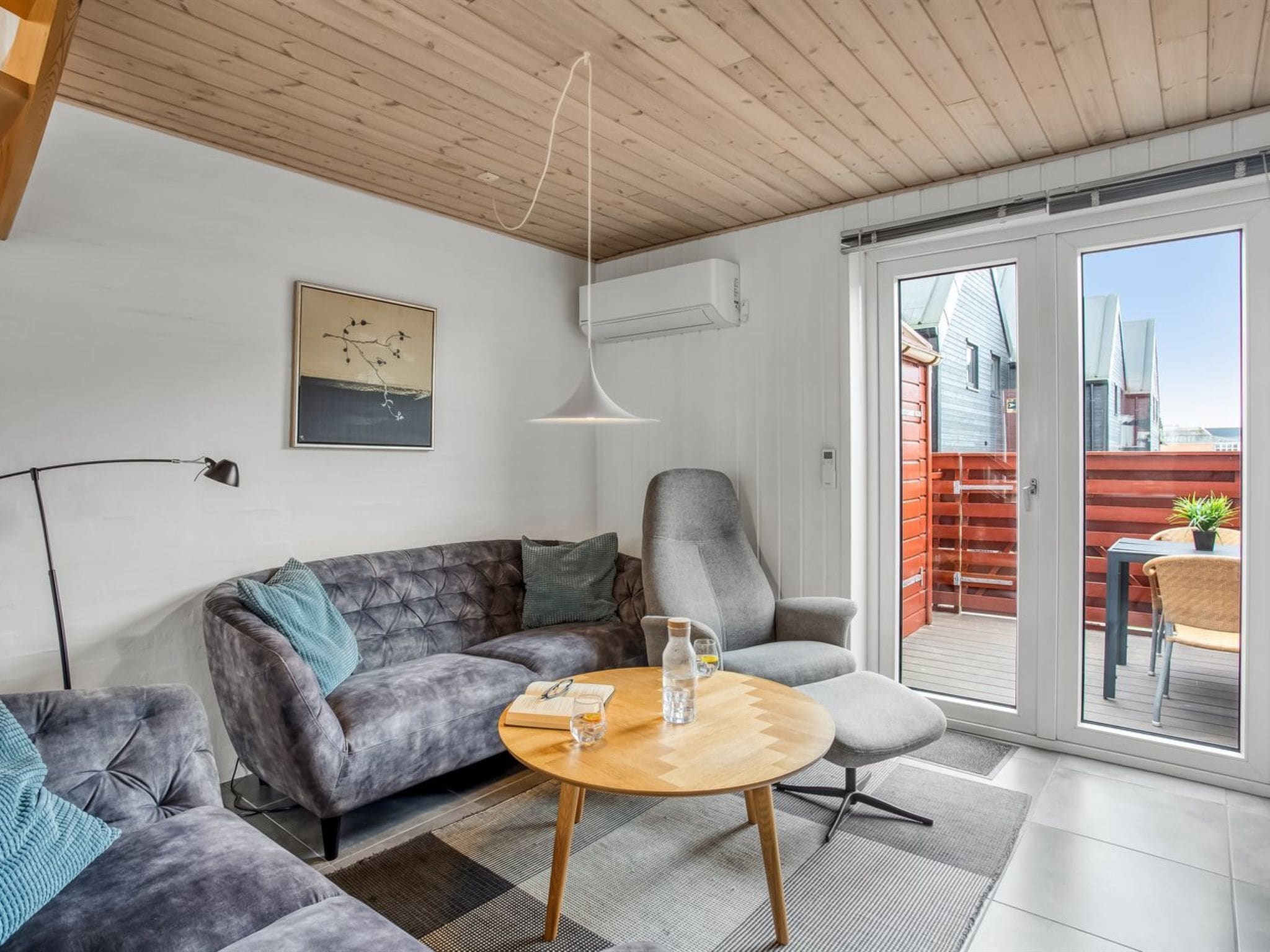 Un saló modern amb un sofà gris, una taula de centre de fusta rodona i un butac armchair gris. Hi ha una finestra gran amb vistes a un edifici vermell.