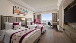 Ein Hotelzimmer mit zwei Betten, einem Schreibtisch, einem Stuhl, einem Fernseher und einem großen Fenster mit Blick auf die Stadt.