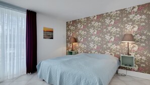 Un dormitorio con cama, dos lámparas de noche, un papel tapiz floral, una ventana con cortinas traslúcidas y un cuadro pequeño en la pared.