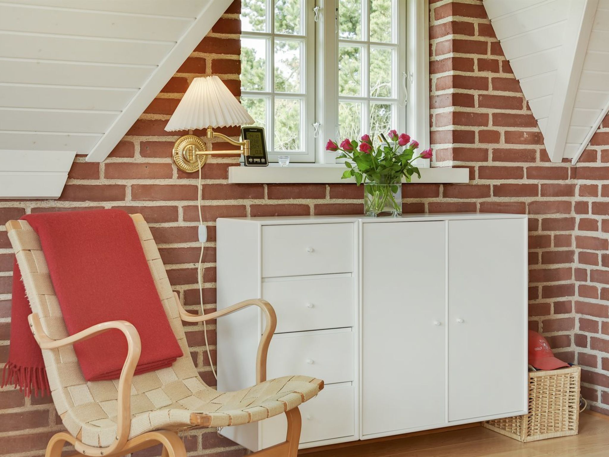 Une pièce chaleureuse avec un mur en briques, une armoire blanche, un fauteuil en osier agrémenté d’un plaid rouge et une fenêtre donnant sur un paysage verdoyant.