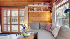 Een gezellig cabin interieur met een houten plafond, een bank met kussens, een eettafel met een fruitschaal en een raam met uitzicht op bomen.