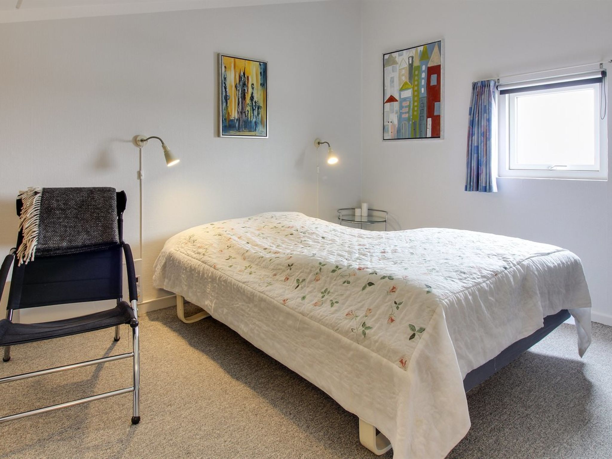 A bedroom with a bed, a chair, a lamp, and two paintings on the wall.