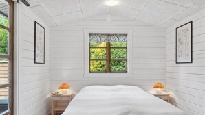 Un dormitorio con una cama, dos mesitas de noche con lámparas, un ventanal con vistas a árboles y una puerta que da al exterior.