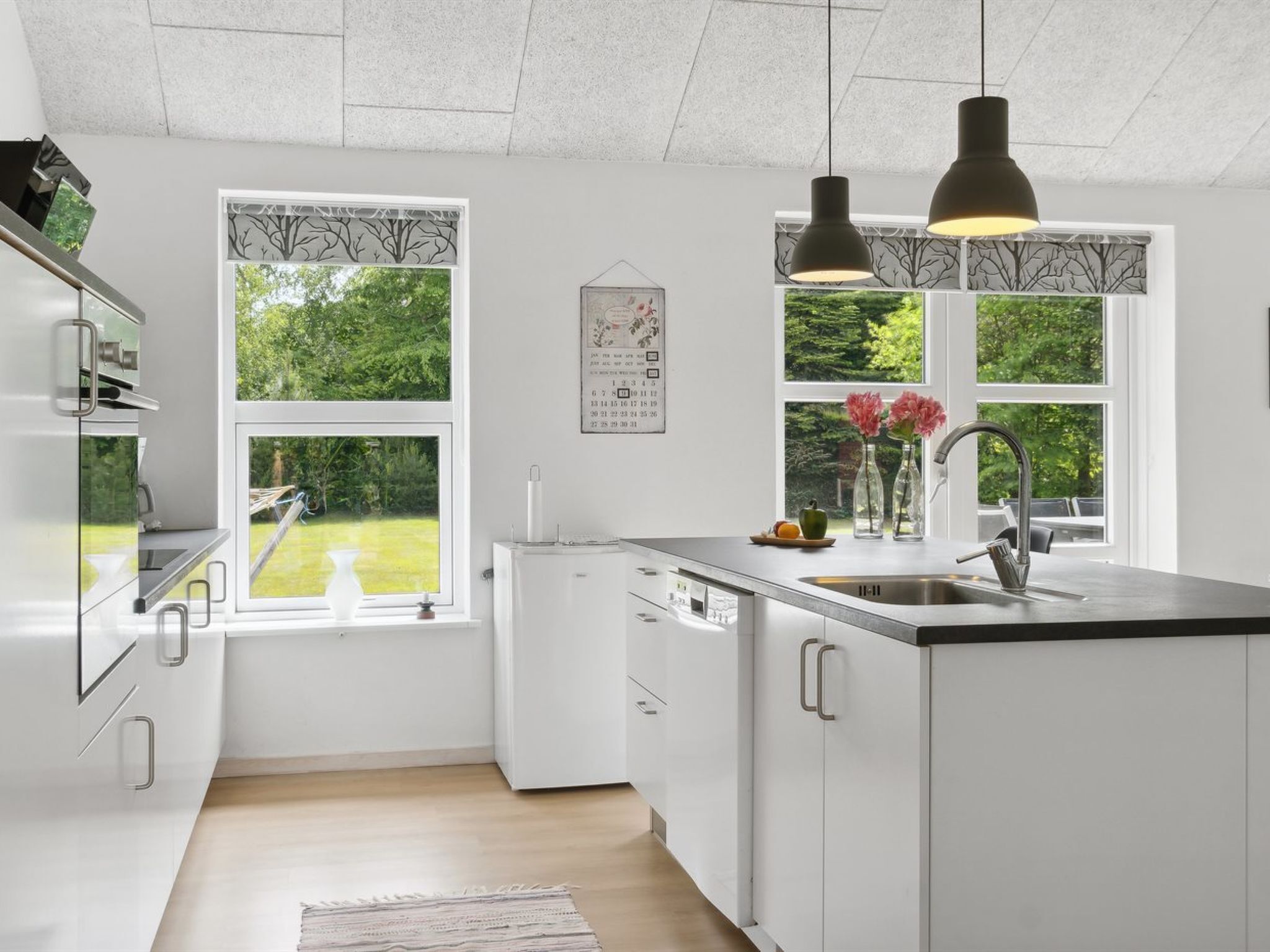A modern kitchen with white cabinetry, a built-in dishwasher, and a large window offering a view of greenery.