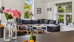 Una sala moderna con un sofá seccional gris, una mesa de comedor de madera y una isla de cocina. Dos ventanas amplias ofrecen luz natural y vistas al follaje exterior.