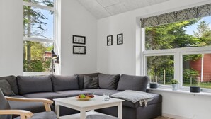Una sala de estar moderna con un sofá seccional gris, una mesa de centro blanca y una mesita auxiliar. En la pared hay cuadros enmarcados y una ventana con vistas a árboles y un edificio rojo.