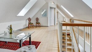Ruime zolderkamer met hoge plafonds, dakramen, een houten trap en een glazen tafel met een rood kleed.