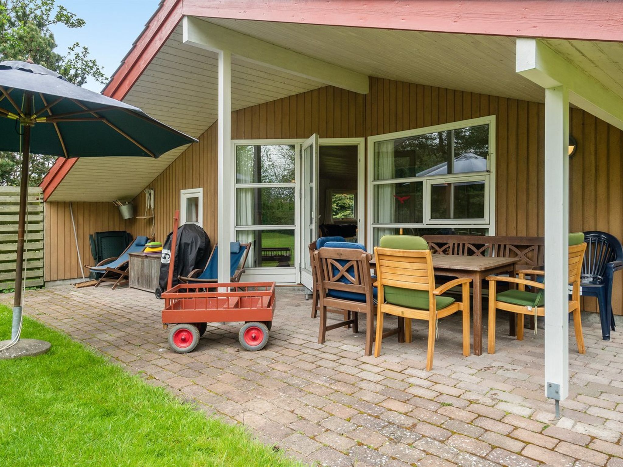 Een overdekt terras met een houten tafel en stoelen, een klein karretje en een parasol.