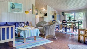 Et hyggeligt rum med trægulv, et stribet tæppe, en blå sofa, fletstole, et spisebord med stole og et køkkenområde i baggrunden.