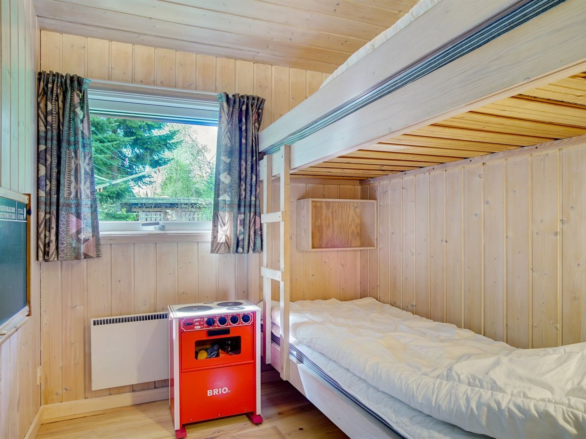 A compact room with a bunk bed, a small kitchenette with a red stove, a window with curtains, and a view of greenery outside.