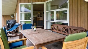 En udendørs terrasse med et træbord og stole, grill og udsigt til indretningen gennem store vinduer.