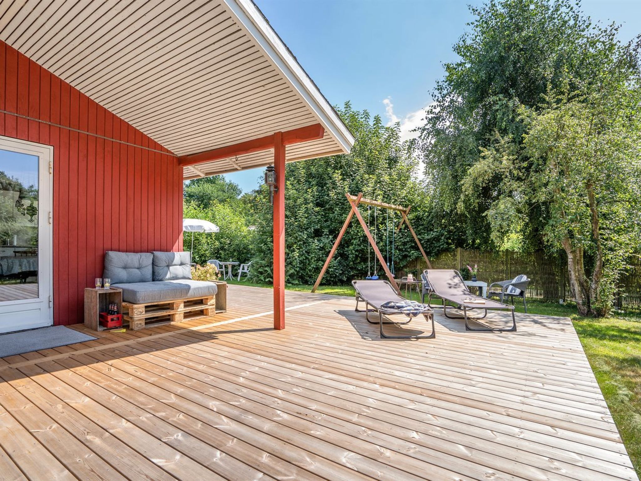 En træterrasse med en rød bygning, gyngestativ og udendørs møbler.