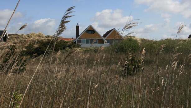 A house with a sloped roof and a chimney, surrounded by tall grass and dunes.